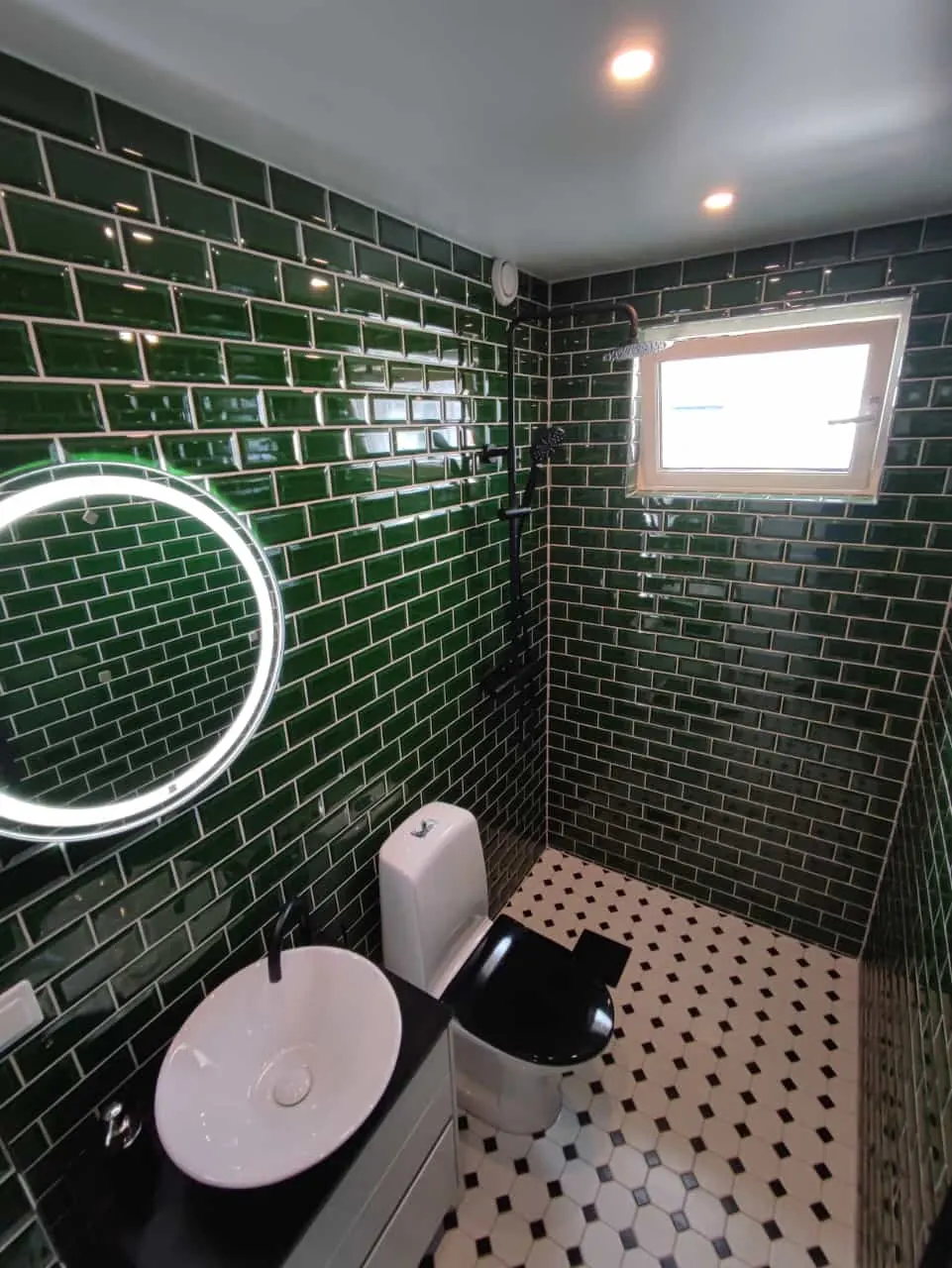 Badrum med grönt fasat kakel, mosaikgolv och rund LED-spegel